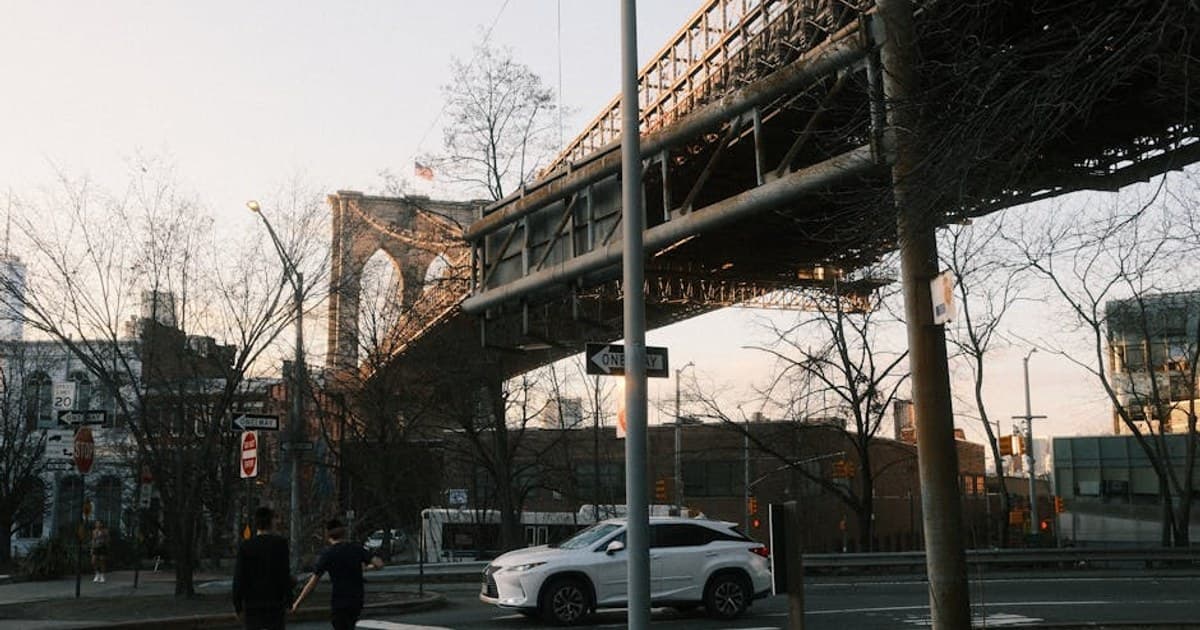 Pedestrians crossing under the Brooklyn Bridge at sunset with urban cityscape.