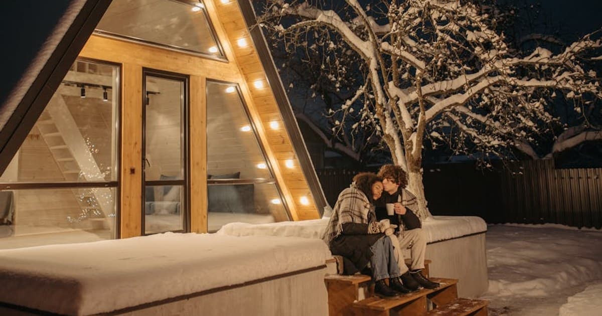 Couple enjoying a warm embrace outside an illuminated A-frame cabin on a snowy winter night.
