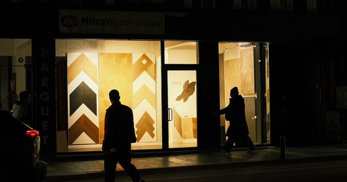 Silhouettes of people walking past an illuminated parquet storefront at night.