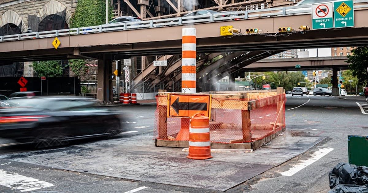 Blurred cars passing beneath Brooklyn Bridge with construction cones.