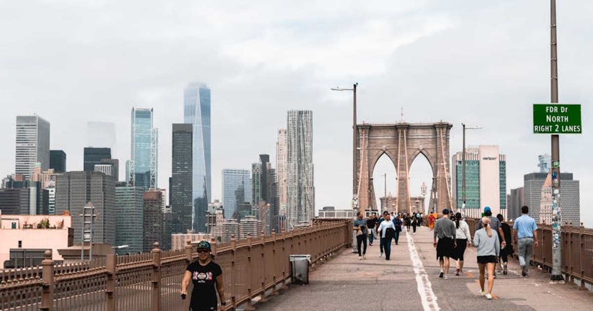People walking on Brooklyn Bridge with Manhattan skyscrapers in the background under cloudy skies.