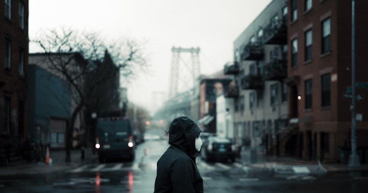 A person in a face mask walks across a Brooklyn street on a foggy day, with the Williamsburg Bridge in the background.