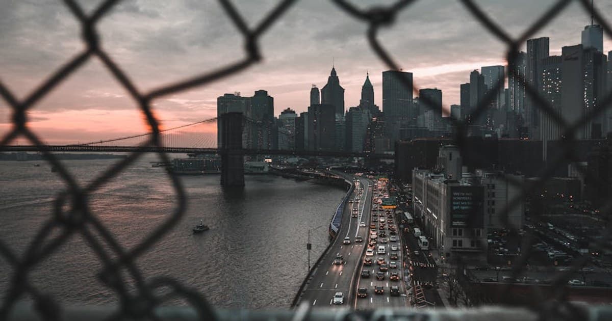 View of New York's skyline and Brooklyn Bridge at dusk through a fence.